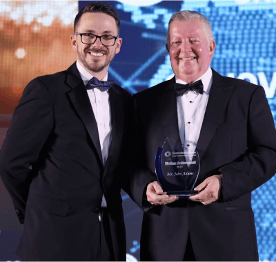 Two smiling men in formal tuxedos, one holding an award trophy, on stage at a maritime industry event